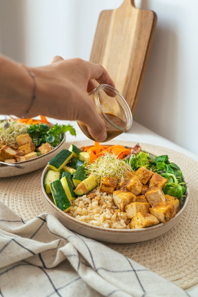 main qui verse une sauce au soja et à la cacahuète sur une assiette de tofu riz et légumes